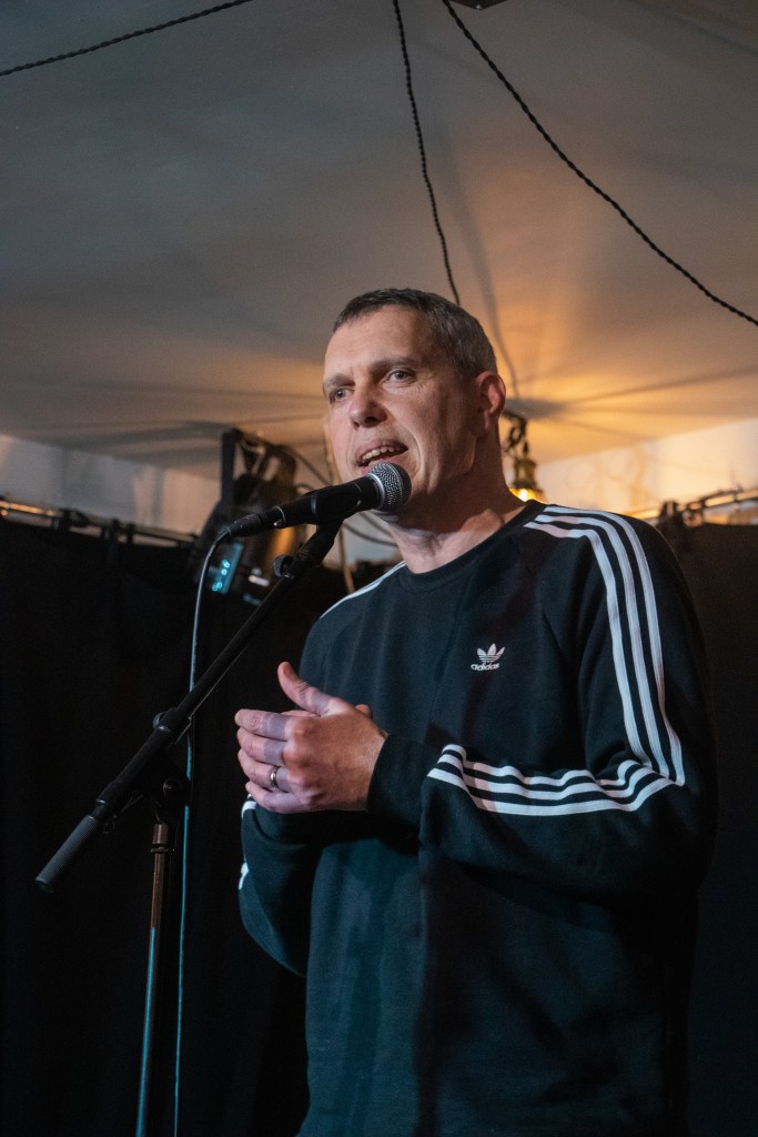 Richard Boggie performing poetry in front of a microphone, wearing a black Addidas jumper